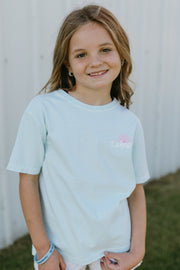 Young girl wearing a light mint t-shirt and white shorts standing in front of a white building.