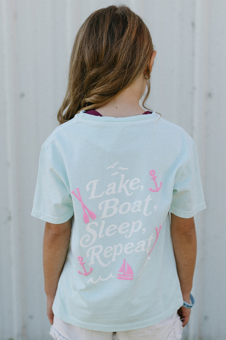 Girl wearing a light mint t-shirt with text and graphics, standing on grass against a white building.