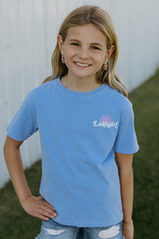 Young girl wearing a peri t-shirt with lakegirl text, standing in front of a white building.