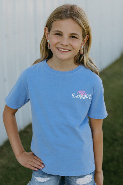 Young girl wearing a peri t-shirt with lakegirl text, standing in front of a white building.