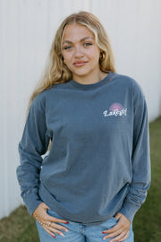 Woman wearing an indigo long-sleeve shirt with a logo, standing in front of a white wall.
