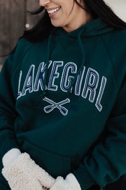 Person wearing a green 'Lake Girl' hoodie with a wooden cabin background
