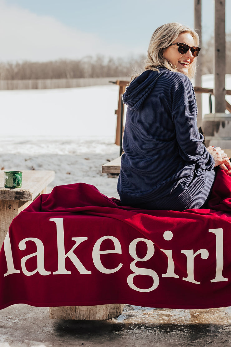 Woman sitting on a bench with a 'Lakegirl' blanket in a snowy outdoor setting
