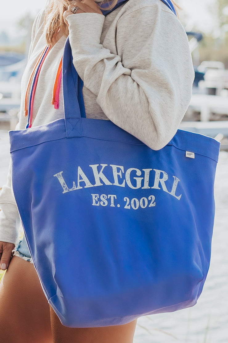 Person holding a peri tote bag with 'LAKEGIRL EST. 2002' text by a waterfront.